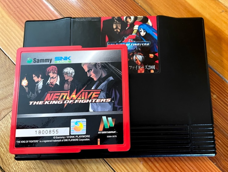 An Atomiswave cart (King of Fighters Neowave) on top of a Neo Geo cart (King of Fighters 2001), showing that one is much larger than the other