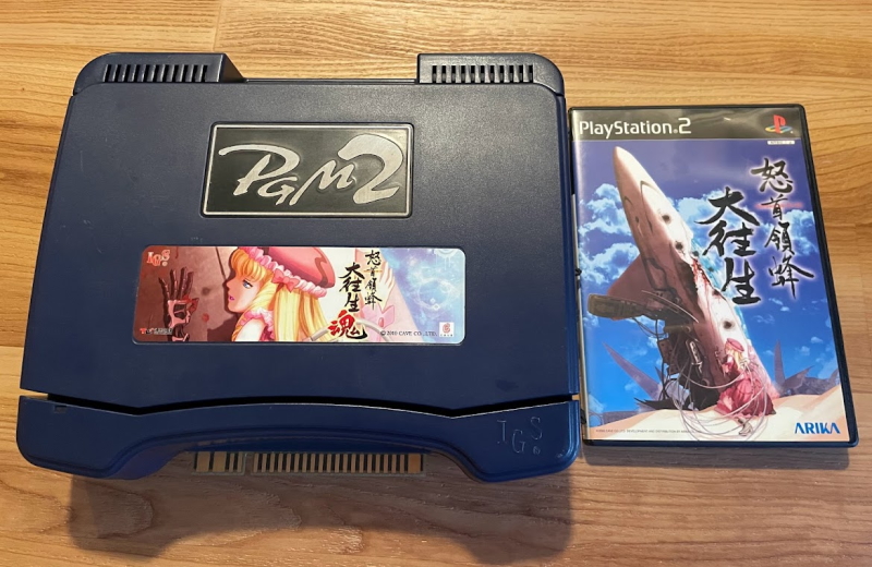 A PGM2 next to a playstation 2 box. The PGM2 is in a curvy blue plastic case, and its label is a cropped version of the playstion 2 game's box art. The box art shows a damaged robot girl, her robotic hand red with the blood of her pilot