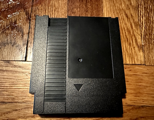 An unlabeled black NES cartridge on a hardwood floor. There is a stain in the corner